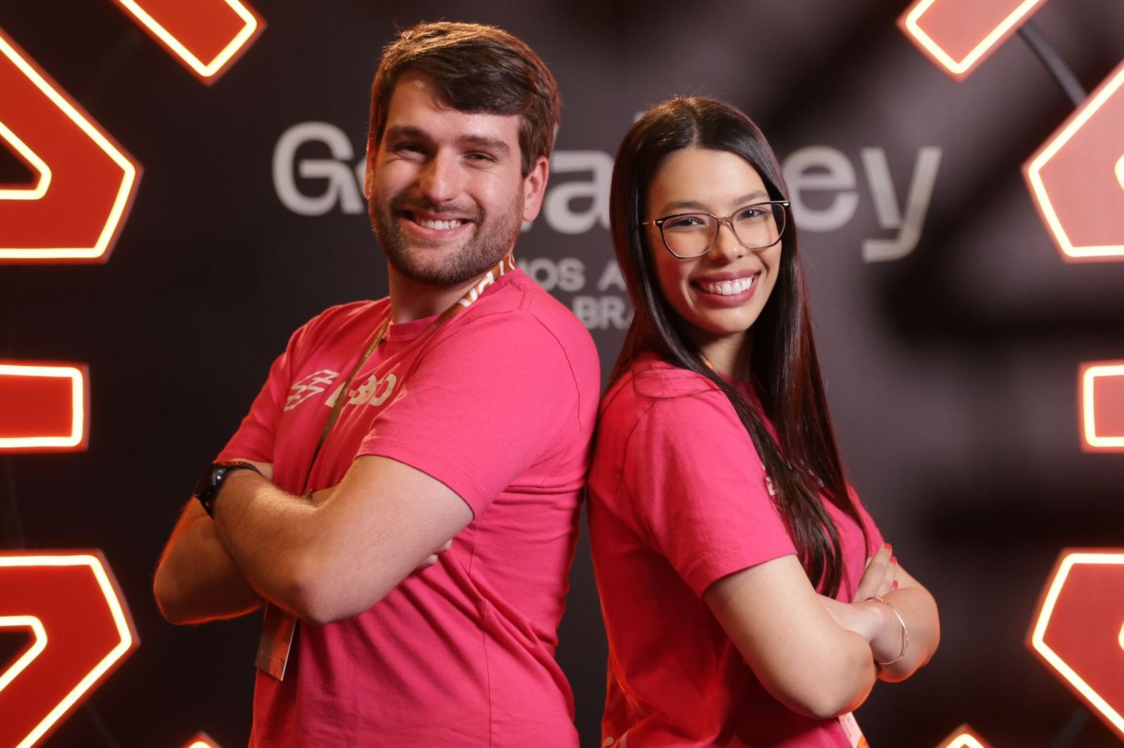 Imagem com os fundadores da Lazi, um homem branco com cabelo castanho claro e barba. Uma mulher com cbaelo castanho escuro longo e óculos preto. Ambos de costas um para o outro com braços cruzados e camiseta rosa.