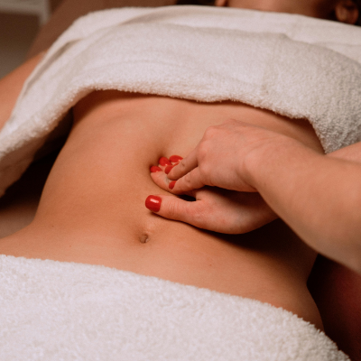 Pessoa deitada em uma maca coberta por toalhas brancas, recebendo massagem abdominal de um profissional durante um procedimento estético ou de bem-estar.